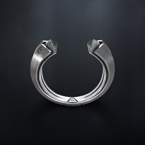 May include: A silver-colored ring with a unique open design. The ring features geometric shapes at the ends and a series of parallel bands forming the main body. The ring is set against a dark background, highlighting its metallic sheen.