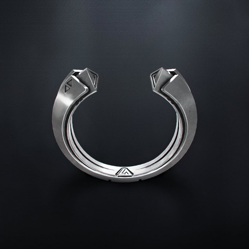 May include: A silver-colored ring with a unique open design. The ring features geometric shapes at the ends and a series of parallel bands forming the main body. The ring is set against a dark background, highlighting its metallic sheen.