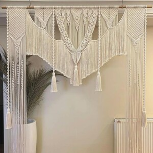 Op de afbeelding: Een grote macramé wandhanger met een geometrisch ontwerp. De hanger is gemaakt van natuurlijk katoenen koord en heeft een franje rand. Hij is opgehangen aan een muur met een houten stok.
