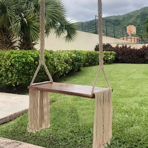 Op de afbeelding: Een houten schommelstoel met een natuurlijke houten afwerking hangt aan twee dikke, witte touwen met kwastjes. De schommel is opgehangen in een grasachtige achtertuin met een weelderig groen gazon.