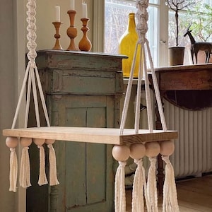 Op de afbeelding: Een houten plank met witte kwastjes hangt aan een macramé touw. De plank is aan het plafond gehangen en heeft houten kralen aan de uiteinden. De plank bevindt zich in een kamer met een vintage dressoir en een raam.