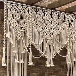 Pode incluir: Uma tapeçaria de macramé branca com um padrão geométrico e franjas. A tapeçaria é feita de cordão de algodão e mede aproximadamente 90 cm de largura e 60 cm de altura.