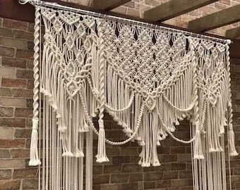 May include: A white macrame wall hanging with a geometric pattern and tassels. The hanging is made of cotton cord and is approximately 3 feet wide and 2 feet tall.