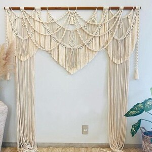 Op de afbeelding: Crèmekleurige macramé wandhanger met een houten staaf. Het ingewikkelde ontwerp heeft geometrische patronen, watervalfranjes en decoratieve kwastjes. Dit textiel in bohemienstijl voegt textuur en warmte toe aan elke binnenruimte.