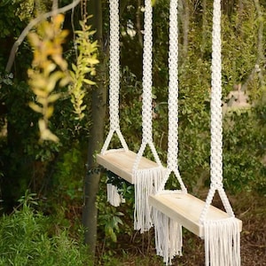 May include: Two wooden shelves with white macrame rope hanging from a tree branch. The shelves are suspended by the rope and have a fringe detail.