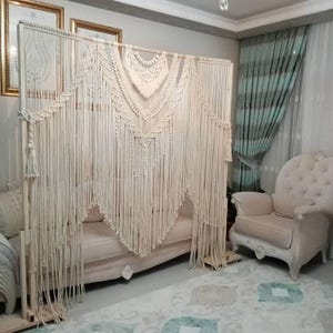 Op de afbeelding: Een witte macramé wandhanger in boho-stijl. De hanger is gemaakt van katoenen koord en heeft verschillende knopen en patronen. Hij hangt aan een houten frame en wordt getoond in een woonkameromgeving.