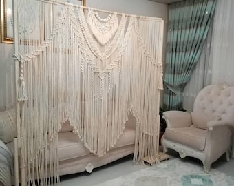 May include: A white macrame wall hanging with a bohemian style. The hanging is made of cotton cord and features a variety of knots and patterns. It is hung on a wooden frame and is displayed in a living room setting.