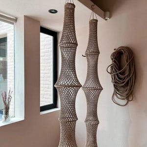 May include: Two beige macrame pendant lights hanging from the ceiling. The lights have a cylindrical shape with a flared base and top, and a woven net design. A coil of rope hangs on the wall to the right.