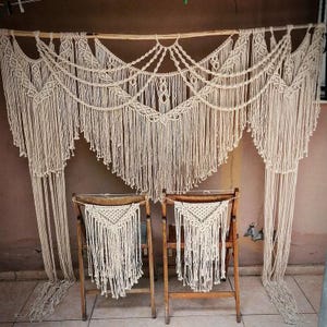 May include: Cream-colored macrame wall hanging with intricate knotting patterns, suspended from a wooden rod. Two wooden chairs with macrame backrests are positioned below. The design features cascading fringe and geometric shapes, creating a bohemian aesthetic.