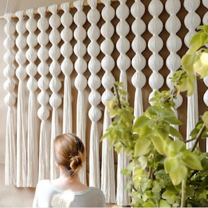 May include: A white macrame wall hanging with a series of large, round knots and long tassels. The wall hanging is made of natural cotton cord and has a bohemian style.