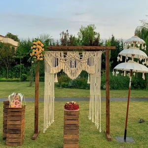 May include: An outdoor wedding ceremony setup featuring a wooden arch with macrame detailing, a tiered white umbrella, and wooden crates. A cake and floral arrangements are displayed on the crates, set on a grassy lawn.