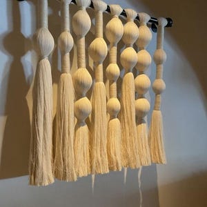 Könnte beinhalten: Ein Wandbehang aus Naturfaserseil mit einer Reihe von geknoteten Kugeln und Quasten. Der Behang ist cremefarben und an einer schwarzen Metallstange befestigt.