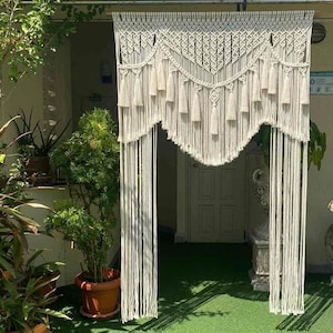 May include: Cream macrame door curtain with a scalloped top edge and hanging tassels. The curtain is made of woven cotton and hangs in front of a doorway. The design includes intricate knotting patterns and long vertical strands.