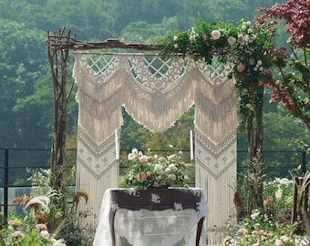 May include: A rustic wedding ceremony arch decorated with a macrame tapestry and greenery. A small table with a white lace tablecloth sits in front of the arch, adorned with a bouquet of white and pink flowers.