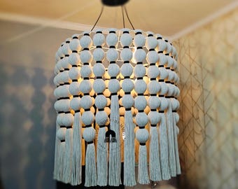 Puede incluir: Una lámpara de araña de macramé blanca con detalles negros y borlas. La lámpara de araña está hecha de una serie de bolas tejidas con una banda negra alrededor de cada bola.