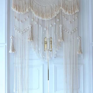 Op de afbeelding: Crèmekleurig macramé deurgordijn. Het gordijn heeft een geweven bovenkant met een geschulpte rand, kwastjes en hangende strengen. Decoratieve bollen en geometrische patronen voegen visuele interesse toe aan dit interieur in bohemien-stijl.