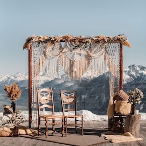 Op de afbeelding: Een rustieke opstelling voor een huwelijksceremonie in de open lucht met twee houten stoelen tegenover een macramé-achtergrond. De achtergrond is versierd met gedroogde bloemen en pampasgras. De scène speelt zich af tegen een achtergrond van besneeuwde bergen en een heldere blauwe lucht.