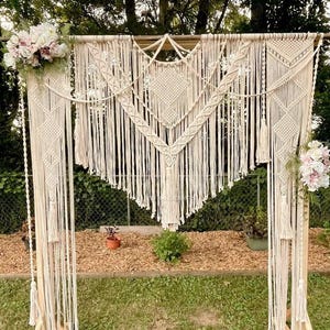 Op de afbeelding: Een witte macramé bruiloft boog met een houten frame, versierd met bloemen. De boog is gemaakt van verschillende knopen en patronen, en hangt aan een houten balk. De bloemen zijn wit en roze, en zijn in een tros aan de bovenkant van de boog gerangschikt.
