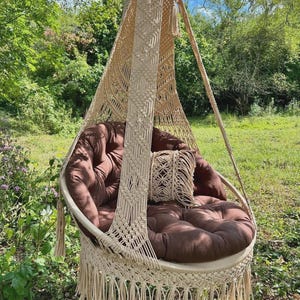 May include: A beige macrame hammock chair with fringe hanging from a tree branch. The chair has a brown cushion and a smaller beige pillow.