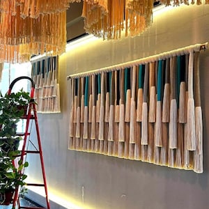 Könnte beinhalten: Ein großer Wandbehang mit natürlicher, böhmischer Ästhetik. Er zeigt vertikale Stränge in Beige-, Türkis- und Brauntönen, die einen strukturierten, geschichteten Effekt erzeugen. Das Stück ist an einer Holzstange befestigt und verleiht ihm einen rustikalen Touch. Der Raum hat auch Hängedekorationen.