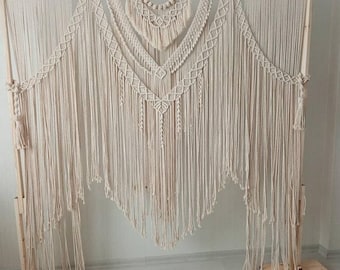 May include: A cream-coloured macrame wall hanging with detailed geometric patterns and fringe. The textile art piece is displayed on a wooden frame, showcasing a bohemian style. The design features a central V-shape and a series of horizontal decorative elements.
