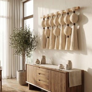 Op de afbeelding: Een beige macramé wandkleed met een houten staaf en decoratieve kwasten. Het wandkleed hangt boven een houten dressoir met een neutrale loper en diverse keramische vazen. Links van het dressoir staat een olijfboom in een pot.