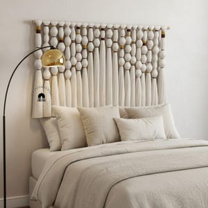 Könnte beinhalten: Cremefarbenes Makramee-Kopfteil mit Holzstange oben. Das Kopfteil hat geknotete, mit Perlen besetzte und gefranste Elemente. Das Bett ist mit cremefarbener Bettwäsche und Kissen ausgestattet. Links ist eine goldene Lampe.