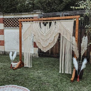 Op de afbeelding: Een houten boog versierd met een groot macramé-wandkleed, perfect voor een bruiloft of een speciale gelegenheid. De boog is buiten opgesteld in een grasveld met gedroogd pampasgras aan beide kanten.