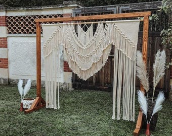 May include: A wooden archway decorated with a large macrame tapestry, perfect for a wedding or special event. The archway is set up outdoors in a grassy area with dried pampas grass on either side.