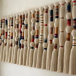 May include: A large, wall-mounted textile art piece. It features multiple vertical strands of off-white, cylindrical shapes with colourful bands of red, blue, brown, and gold. The bottom of each strand has a long, tassel-like fringe.