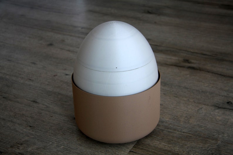 K&ouml;nnte beinhalten: Ein wei&szlig;er, eif&ouml;rmiger Beh&auml;lter mit einem braunen Deckel. Der Beh&auml;lter ist aus einem glatten, matten Material gefertigt.