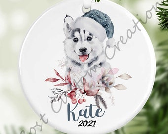 May include: White ceramic Christmas ornament featuring a watercolor illustration of a husky puppy wearing a Santa hat, surrounded by red berries and greenery. The ornament is personalized with the name "Kate" and the year "2021".