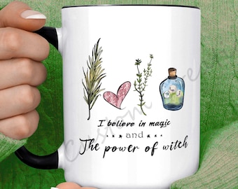 May include: White ceramic mug with a black rim and handle. The mug features a watercolor illustration of a heart, herbs, and a bottle with a cork. The text reads "I believe in magic and the power of witch".