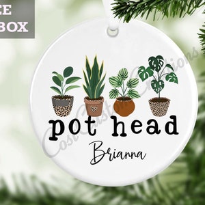 Pode incluir: Ornamento de cerâmica branco com uma ilustração verde, marrom e branca de plantas em vaso. O texto "pot head" e o nome "Brianna" estão impressos no ornamento.