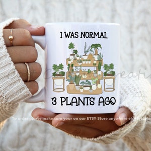 May include: White ceramic mug with a black and white graphic of a shelf with many potted plants. The text "I was normal 3 plants ago" is printed on the mug.