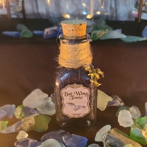 May include: A small glass bottle with a cork stopper and a brown twine tied around the neck. The bottle is filled with a glittery purple liquid and has a label that reads "Bat Wing Tonic" with a small bat illustration.