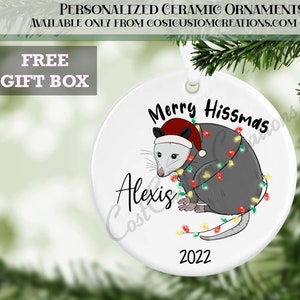 May include: A white ceramic Christmas bauble featuring a grey opossum wearing a red Santa hat and surrounded by colourful Christmas lights. The bauble reads "Merry Hissmas" and "Alexis" in black text. The year "2022" is printed in black at the bottom of the bauble.