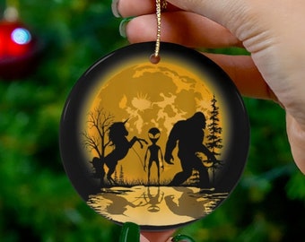May include: A round, black and gold ornament featuring a silhouette of a unicorn, alien, and Bigfoot in front of a full moon. The ornament has a gold string for hanging and is held against a Christmas tree background.