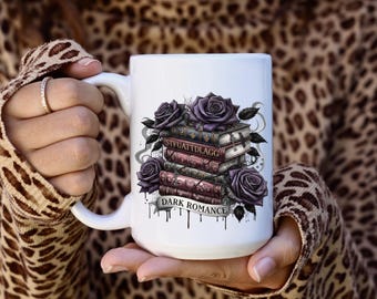 May include: White ceramic mug featuring a stack of books with the text "STFUATDLAGG" and "DARK ROMANCE". The design includes purple roses and a banner. The mug is held in a person's hands.