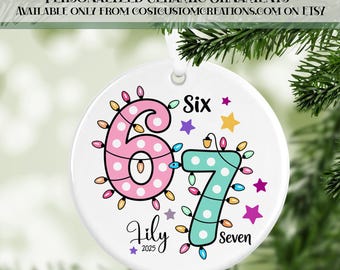 May include: White ceramic ornament with the numbers "67" in pink and teal, decorated with string lights. The ornament includes the name "Lily" and the year "2025". The text "Six" and "Seven" are also included.