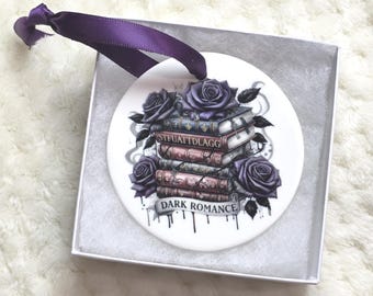 May include: A white ceramic ornament featuring a stack of books and purple roses. The ornament has the words "DARK ROMANCE" and a purple ribbon for hanging. The ornament is in a white gift box.