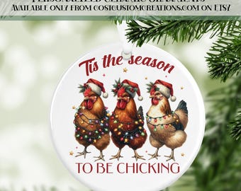 May include: A white ceramic Christmas ornament featuring three chickens wearing Santa hats and wrapped in string lights. The text "Tis the season to be chickin" is written in red. The ornament is hanging from a white string.