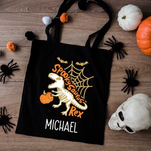Puede incluir: Bolsa de lona negra con un gráfico de dinosaurio esqueleto blanco y el texto "Spooky Saurus Rex" y "MICHAEL". La bolsa está rodeada de pequeñas calabazas blancas y naranjas, arañas negras y una calavera blanca.
