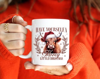 May include: White ceramic mug featuring a Highland cow wearing a Santa hat, with the text "Have Yourself a Mooey Little Christmas." The design includes a wreath and snowflakes. A festive holiday-themed mug.