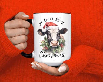 May include: White ceramic mug with a black handle and rim. The mug features a cow wearing a Santa hat and a wreath, with the words "MOOEY Christmas" in a festive font. The mug is held by a person wearing a red sweater.