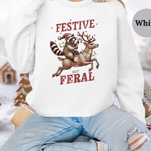 Op de afbeelding: Wit sweatshirt met een feestelijke wasbeer die op een rendier met kerstverlichting rijdt. De tekst luidt "Festive But Feral".