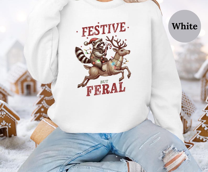 Op de afbeelding: Wit sweatshirt met een feestelijke wasbeer die op een rendier met kerstverlichting rijdt. De tekst luidt "Festive But Feral".