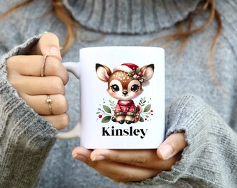 May include: White ceramic mug featuring a cartoon deer wearing a Santa hat and a red and green sweater. The deer is surrounded by green leaves and berries. The name "Kinsley" is printed in black below the deer.