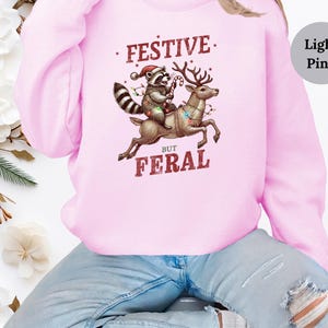 Op de afbeelding: Een lichtroze sweatshirt met een afbeelding van een wasbeer die op een rendier rijdt met kerstverlichting. De tekst op het sweatshirt luidt "Festive But Feral".