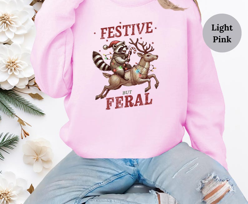 Op de afbeelding: Een lichtroze sweatshirt met een afbeelding van een wasbeer die op een rendier rijdt met kerstverlichting. De tekst op het sweatshirt luidt "Festive But Feral".